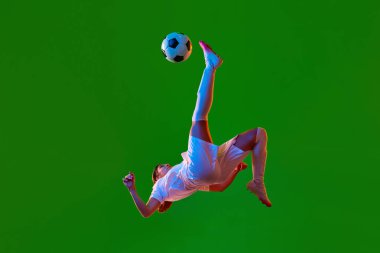 Genç bir kızın dinamik görüntüsü, hareketli bir futbolcu, topa tekme atıyor ve neon ışıkta yeşil zemine doğru düşüyor. Profesyonel spor kavramı, rekabet, oyun, eğitim, eylem, reklam