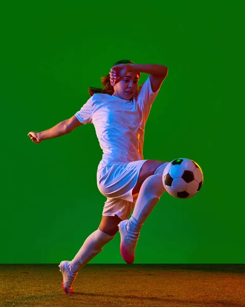 Dynamic image of young woman, football player in motion, kicking ball in jump with knee against green background in neon light. Concept of professional sport, competition, game, training, action, ad