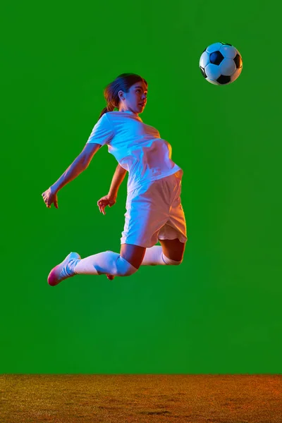 Young woman, football player in motion, kicking ball with breast against green studio background in neon light. Concept of professional sport, competition, game, training, youth, action, ad