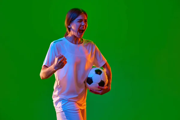 Winner. Competitive young girl, football player posing with ball against green studio background in neon light. Success. Concept of professional sport, competition, game, training, youth, action, ad