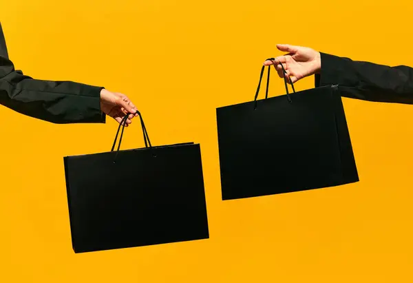 Sale offer. Black Friday. Shopping discount. Closeup of two womans hands holding purchase black bags isolated on bright yellow background. concept of fashion, beauty, salesperson. copy space. ad