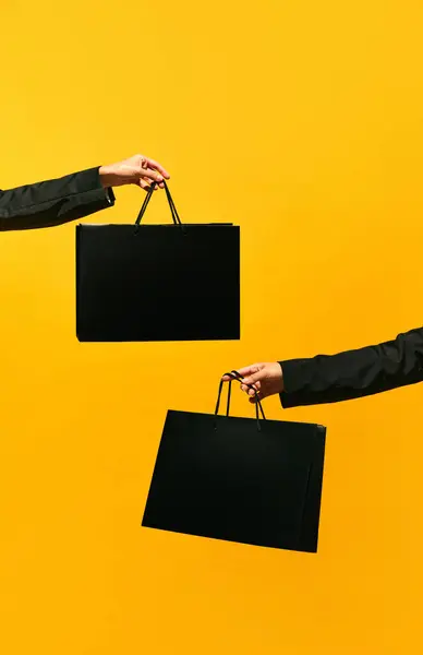 Sale offer. Black Friday. Shopping delivery. Closeup of two womans hands holding purchase black bags isolated on bright yellow background. concept of fashion, beauty, salesperson. copy space. ad