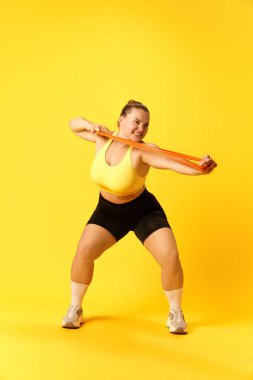 Kendine güvenen, eşofmanlı, spor ayakkabılı ve sarı arka planda spor lastiği takan bir kadının tam boy portresi. Spor, hobi, sağlık, yaşam tarzı, egzersiz kavramı