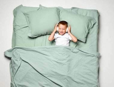 Sırtüstü uzanmış, kulaklıkla müzik dinleyen küçük bir çocuğun portresi. Hoş çocuk, büyük bir nane yatağında bir çocuk. Duygu kavramı, cihazlar, sabah, uyku sağlığı, uyanma..