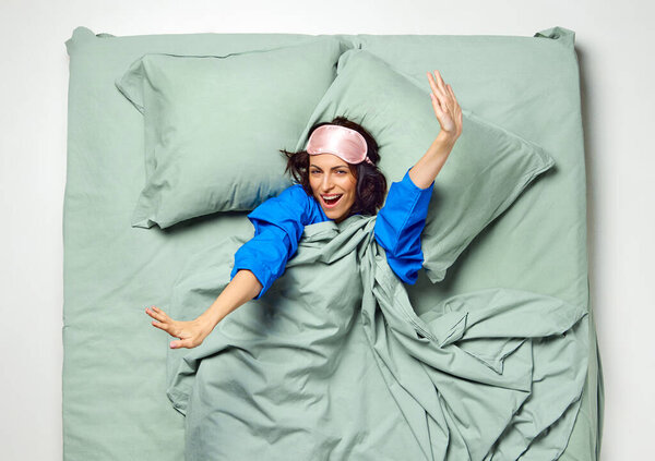 Young beautiful woman with sleeping mask covered with blanket raising up stretching arms for wake up feels. Enough night sleep. early morning. Concept of recreation, rest, bedtime, hotel bedroom. ad