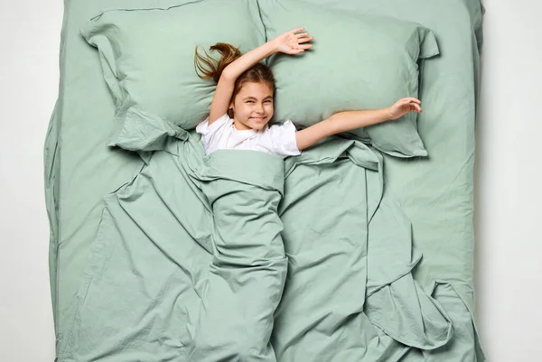 トップビュー カメラを見ている小さな美しい女の子が手を挙げています 幸せな陽気な子供 大きなミントベッドでリラックスする子供 睡眠ウェルネス 人間のタッチ 目覚め ハッピー 就寝の概念 アドベンチャー — ストック写真