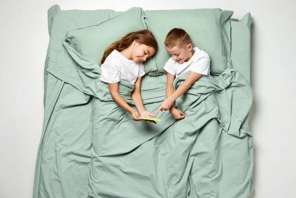 뒷면에 누워있는 소녀와 소년의 초상화는 전화로보고 만화를보고 담요로 사랑스러운 침대에 — 스톡 사진
