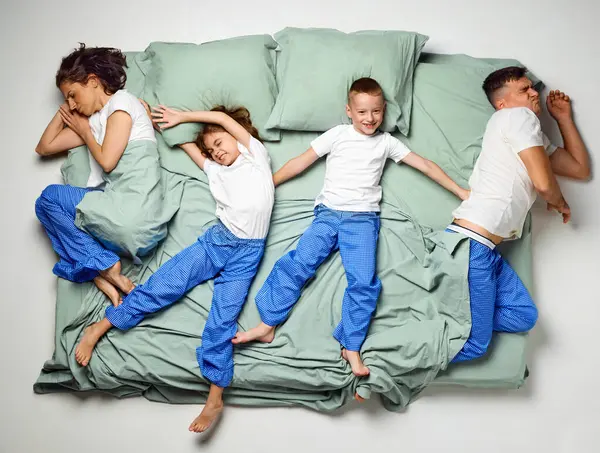 Top view of people covered with blanket lying in bed. Children lying crossed bed and parents on edge of bed. Family sleeping. Concept of sleep wellness, family time, awake, love, morning. ad