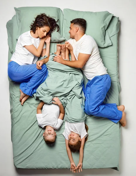 Top view portrait of people covered with blanket lying in bed. Children lying crossed bed and parents on edge of bed. Family sleeping. Enjoying being together. Concept of sleep wellness, family time.