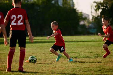 Çocukların tam boy fotoğrafı, spor üniformalı futbolcuların güneşli bir yaz gününde futbol sahasında topla oynamaları. Spor anlayışı, sağlıklı yaşam tarzı, egzersiz, aktivite, hobi.