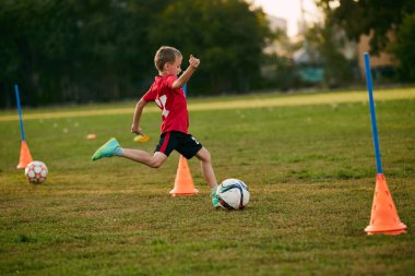 Kırmızı spor üniformalı çocuk futbolcu topa vuruyor, futbol sahasında hareket etmeye hazır olun. Futbol oynuyordum. Çocuklar takım oyunu. Spor yaşam tarzı, aktivite, hobi anlayışı. Ad