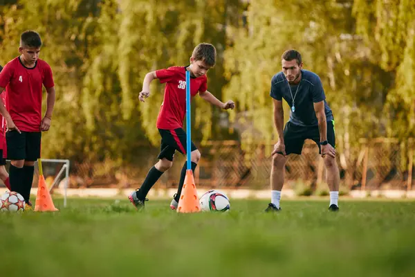 Kids, football players in sport uniform with coach training running with obstacles with ball before match on soccer field. Concept of sport inventory, workout, leisure activity, healthy lifestyle. Ad
