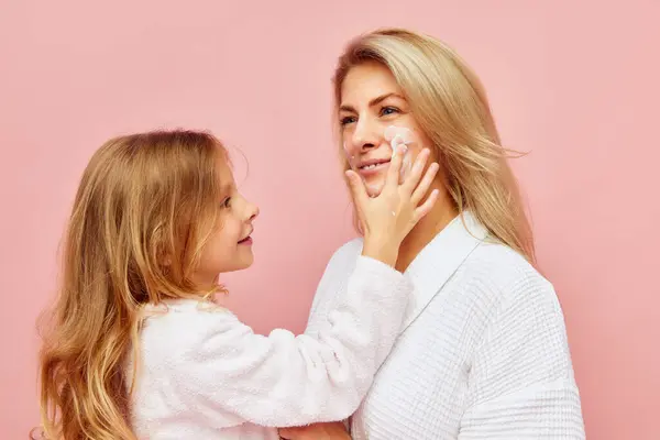 Evening hygiene. Smiling beautiful daughter, girl apply natural cosmetic cream on face, cheeks of young mother, woman over pastel pink background. Concept of fashion, beauty, spa treatment. Ad