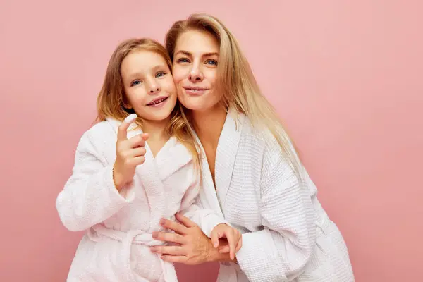 Evening hygiene. Smiling beautiful mother, woman and sweet, lovely, daughter, girl with natural cream on finger over pastel pink background. Concept of fashion, beauty, spa treatment, cosmetic. Ad