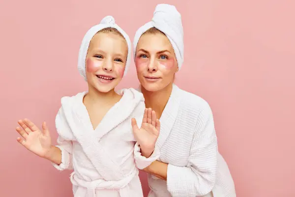 Young attractive woman mother, and cheerful girl, daughter in bathrobes and towels with eyepatches over pastel pink background. Concept of beauty, spa treatment, natural cosmetic, self care. Ad