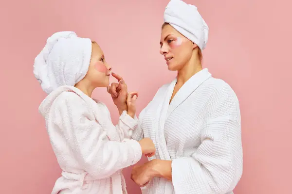 Young attractive woman mother, and girl, daughter in bathrobes and towels with eyepatches doing morning skin care routine over pastel pink background. Concept of beauty, spa, natural cosmetic. Ad