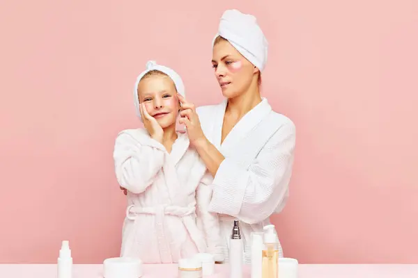Loving, happy mother and daughter in bathrobes and towels using beauty product for skincare, eye patches. Women and child together care of beauty, health. Concept of family spa treatment, cosmetic.