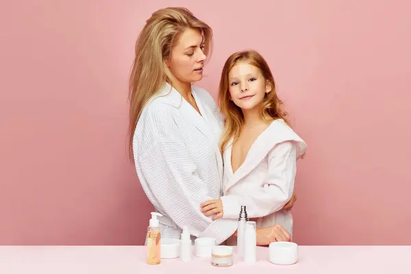 Loving, happy mother and daughter in bathrobes near beauty table. Woman and child together care of beauty, health. Cosmetics and wellness of skin. Concept of family spa treatment, body and self care.