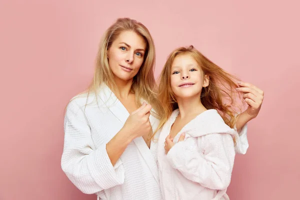 Loving, happy mother and daughter in bathrobes near beauty table looking at camera. Woman and child together care of beauty, health. Cosmetics and wellness of skin. Concept of spa treatment, self care