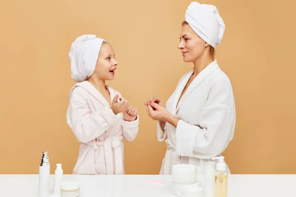 Side view portrait of mother and daughter filing nails in bathroom over beige backgrounds. Woman and kid together care of beauty, health. Concept of spa, wellness, pedicure, manicure, hygiene. ad