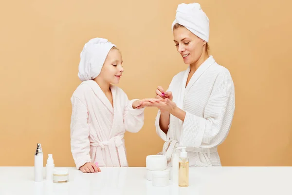 Side view portrait of mother painting nails for daughter in bathroom over beige backgrounds. Woman and kid together care of beauty, health. Concept of spa, wellness, pedicure, manicure, hygiene. ad