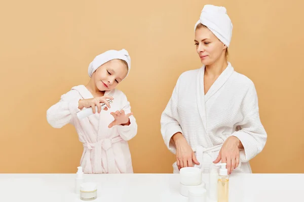 Portrait of lovely mother and daughter apply hand cream, sanitizer, moisturized serum over beige backgrounds. Woman and kid together care of beauty, health. Concept of spa, wellness, hygiene. ad