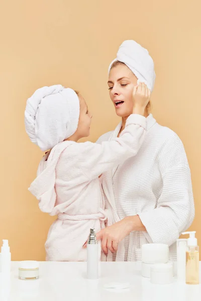 Portrait of cheerful daughter apply cream, moisturized serum to mother face over beige backgrounds. Woman and kid together care of beauty, health. Concept of spa, hygiene, skin care products. ad