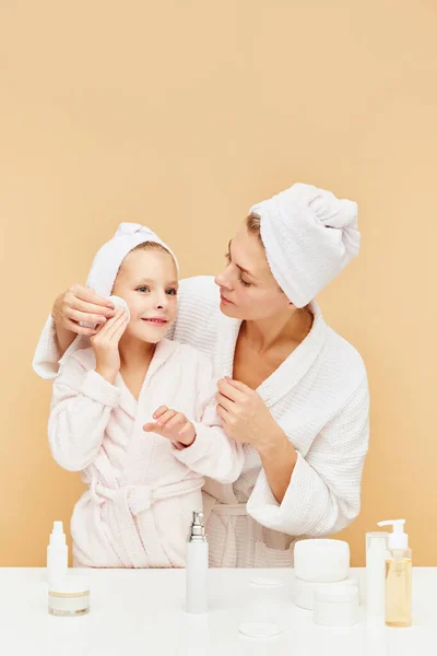 Portrait of cheerful mother teach daughter apply cream, moisturized serum, lotion over beige bathroom backgrounds. Woman and kid together care of beauty, health. Concept of spa, skin care products. ad
