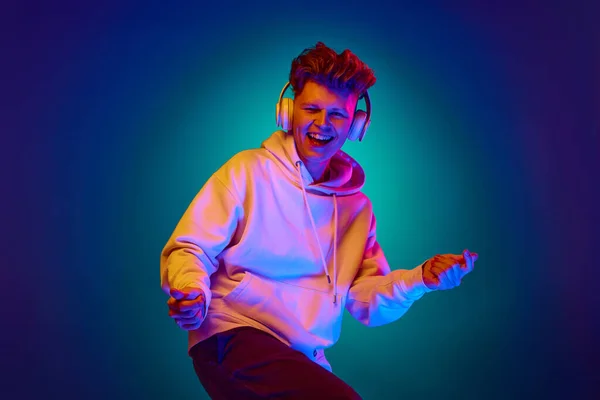 Portrait of attractive young guy, man, dressed white hoodie listening music in headphones and dancing in mixed colors neon filter, light. Concept of human emotions, fashion, style, trends, media. ad