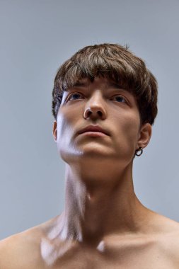 Gri stüdyo arka planına bakan, çıplak omuzlu, esmer, genç bir adamın yakın portresi. Erkek sağlığı, güzellik, moda, vücut ve cilt bakımı, sağlık kavramı. Ad