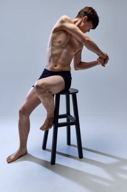 Atletik vücutlu yakışıklı bir adam. Sandalyeye oturmuş bakışlarını kaçırıyor ve gri stüdyo geçmişine karşı ellerini kaldırıyor. Gençlik kavramı, sağlık, doğal güzellik, moda, sağlık, erkeklik.