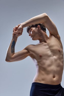 Atletik vücutlu yakışıklı bir adam gözlerini kaçırıyor ve gri stüdyo geçmişine el kaldırıyor. Erkek kaslı vücut. Gençlik kavramı, doğal güzellik, moda, sağlık, erkeklik..