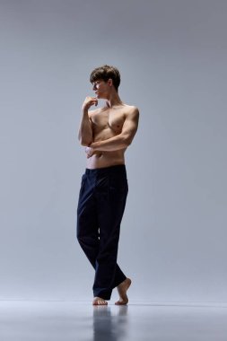 Gri stüdyo arka planında izole edilmiş, yarı çıplak, stoik pozlar veren genç bir adamın tam boy portresi. Erkek kas yapısı geri geldi. Erkek sağlığı, güzellik, moda, vücut ve cilt bakımı, sağlık kavramı. ad