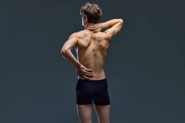 Arkadan bakınca, koyu gri stüdyo arka planında izole edilmiş, boynu ve kaslı sırtını tutan esmer bir adam pozu veriyor. Erkeklerin sağlık, güzellik, moda, sağlık kavramı.