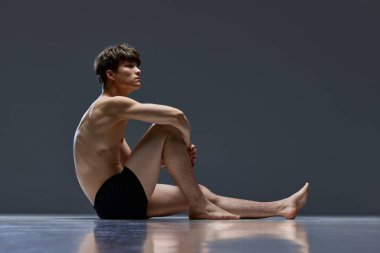 Kaslı vücutlu, iç çamaşırlı, koyu gri stüdyo arka planında oturan genç bir adamın tam boy fotoğrafı. Yan görüş. Erkek sağlığı, doğal güzellik, moda, sağlık kavramı. ad