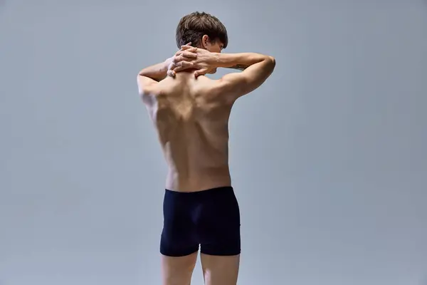 Back view portrait of young handsome man posing shirtless in underwear isolated over grey studio background. Muscular build back. Concept of beauty, fashion, body and skin care, wellness, masculinity.