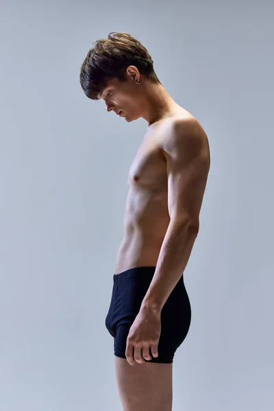 Half-length side view portrait of young man looking down, posing in underwear isolated over grey studio background. Male muscular build body. Concept of beauty, body and skin care, masculinity. ad