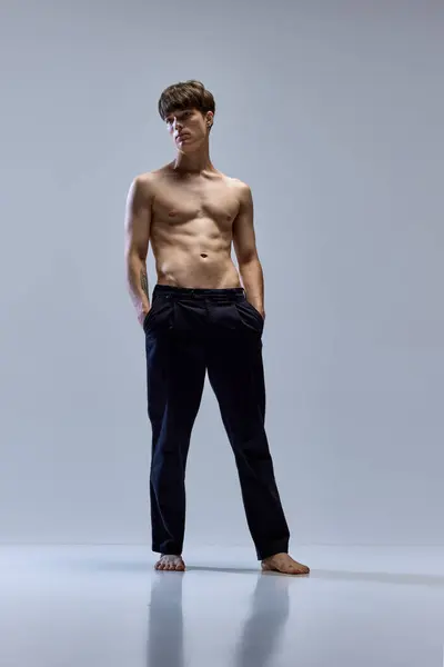 Full-length portrait of young man posing looking away in trousers isolated over grey studio background. Male muscular build body. Concept of natural beauty, body and skin care, masculinity.