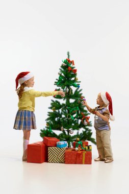 İyi tatiller. Neşeli küçük çocuklar, eski giysiler içinde terli çocuklar oyuncaklarla dekore ediyorlar ve beyaz stüdyo arka planında Noel ağacını selamlıyorlar. Yeni yıl kavramı, çocukluk. Boşluğu kopyala.