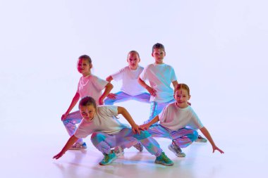 Bir grup gülümseyen tatlı kız, moda kıyafetleri içinde makyaj yapmış çocuklar beyaz stüdyo arka planında neon ışığı altında dans ediyorlar. Hobi kavramı, müzik, hareket, çocukluk. Ad.