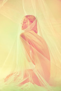 İnce vücutlu çıplak genç kız silueti ıslak ve şeffaf kumaşla kaplı yerde tek başına oturuyor. Retro pastel renkli fotoğraf efekti. Kadın vücudunun doğal güzelliği, kadınlık kavramı.