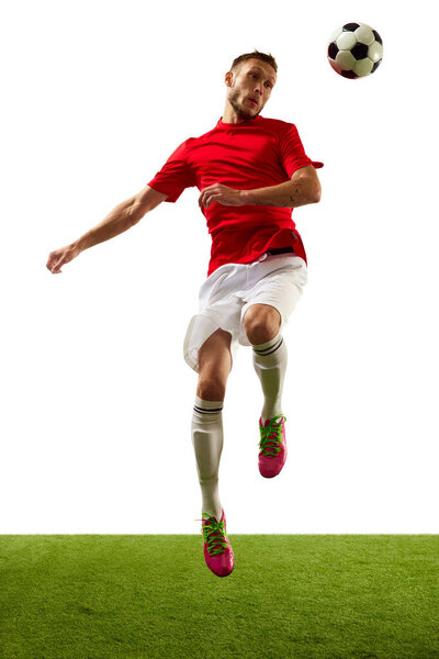Energetic young man, professional player in red sportswear and boots training football tricks against white background with green grass. Concept of game, sport, recreation, active lifestyle.