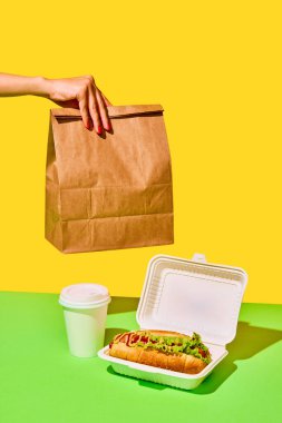 Sosisli, hardallı ve ketçaplı sosisli sandviç ve yeşil-sarı arka planda kağıt torba. Fast food kavramı, sokak yemekleri, teslimat. Metin için boşluk kopyala, ad