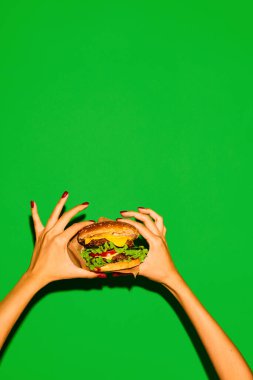 Amerikan yemeği. Elleri, canlı yeşil arka planda geleneksel sıcak hamburger ya da çizburger gibi iştah açıcı. Abur cubur, menü, teslimat, yemek servisi, paket servis. Uzayı kopyala Ad