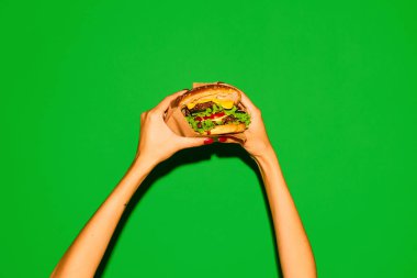 Lezzetli fast-food. Elleri, canlı yeşil arka planda geleneksel sıcak hamburger ya da çizburger gibi iştah açıcı. Abur cubur, menü, teslimat, yemek servisi, paket servis. Uzayı kopyala Ad