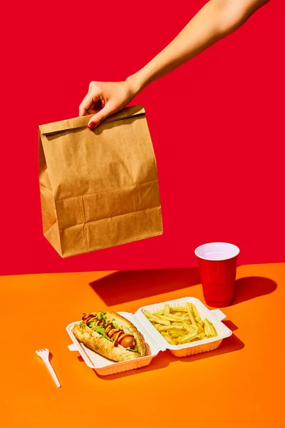 Tasty hotdog with hot sausage served mustard and ketchup and green salad with fried potato and drink against vivid red background. Concept of fast food, street food, menu, catering, take away.