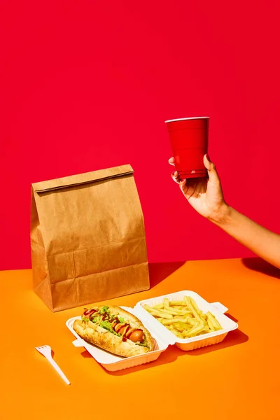 Packing food boxed take away. Tasty hotdog with hot sausage served mustard and ketchup and green salad with fried potato and drink against vivid red background. Concept of fast food, menu, catering.