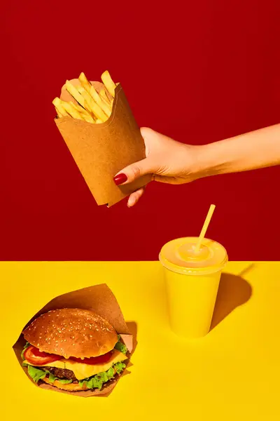 Packing food boxed take away. Appetizing hamburger with beef, sauces and vegetables with soda and fried potato in paper box against vivid red-yellow background. Concept of food, delivery, catering.