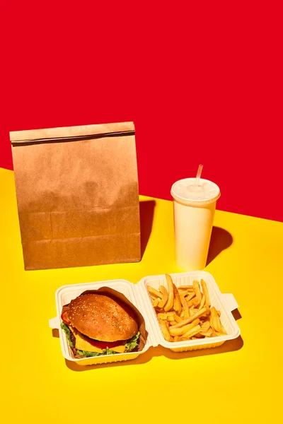 Packing food boxed take away. Hamburger with beef, sauces and vegetables with soda and fried potato in paper boxes against vivid red-yellow background. Concept of junk food, delivery, catering.