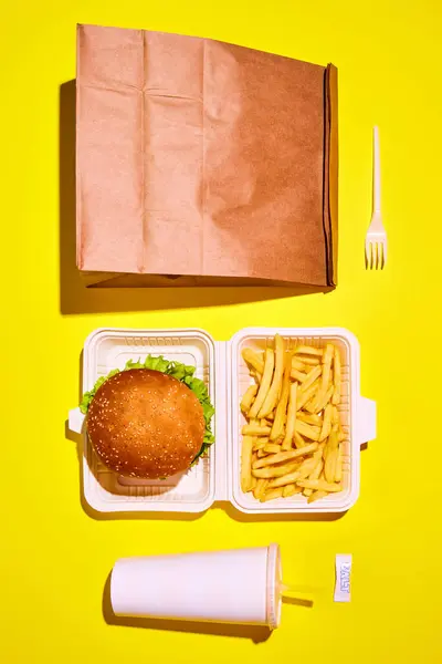 Packing food boxed take away. Top view image of hamburger with beef, sauces and vegetables with soda and fried potato in paper boxes against yellow background. Concept of food, delivery, catering
